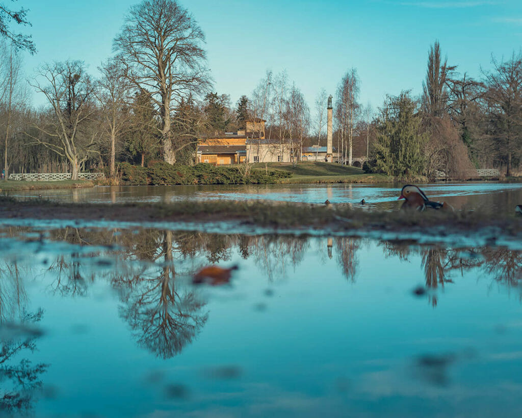 Die Römischen Bäder spiegeln sich in einer Pfütze, dahinter liegt der kleine Teich.